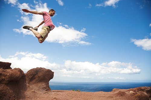 Young fearless hiker jumping off the rock with bent legs on ocean background, Tenerife