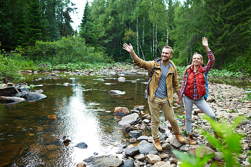 Cheerful hikers waving hands while standing on stones by forest river