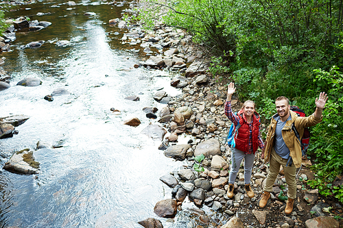 Amorous hikers standing on stones by water and waving hands