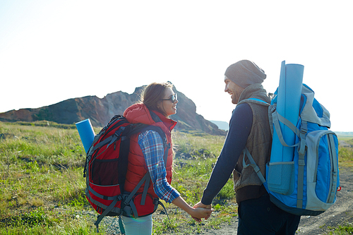 Back view of smiling traveling couple with big backpacks looking happily at each other on wild path in mountains