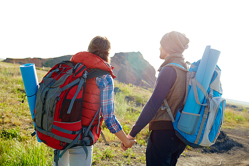 Rear view image of young tourist couple with big backpacks looking away holding hands walking to mountains together