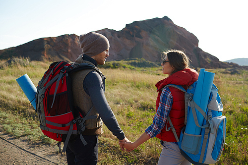 Happy young couple traveling together in mountains: man and woman looking at each other holding hands on hiking path on sunny day