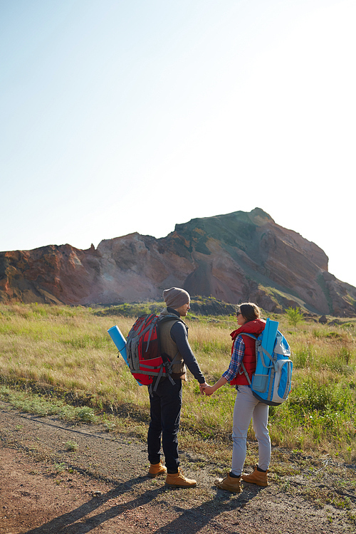 Back view portrait of young couple traveling together in mountains, standing on hiking path with tourist gear and big backpacks holding hands on sunny day