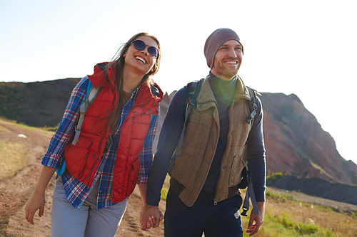 Portrait of smiling young couple wearing active clothes smiling brightly walking on hiking path in mountains together holding hands on sunny day