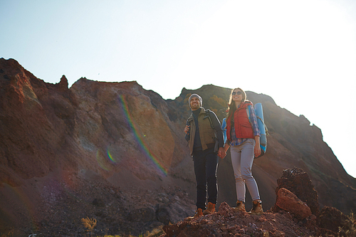 Sunlit image of young traveling couple, man and woman, enjoying view in mountains, standing on top of hill holding hands