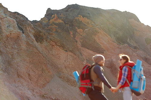 Couple of young travelers, man and woman, hiking in wild nature, man and woman wearing tourist gear with big backpacks standing holding hands against backgrounds of enormous mountain, wide shot with copy space