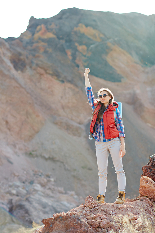 Modern girl in fashionable tourist gear standing on top of peak in mountains raising hand up, inspired and enjoying her freedom