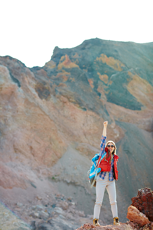 Modern girl in fashionable tourist gear standing on top of peak in mountains raising hand up, inspired and enjoying her freedom