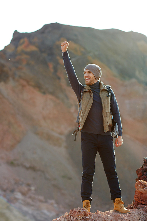 Motivational image of young handsome traveler wearing fashionable clothes standing on top of peak in mountains raising hand up in excitement and smiling, enjoying his freedom