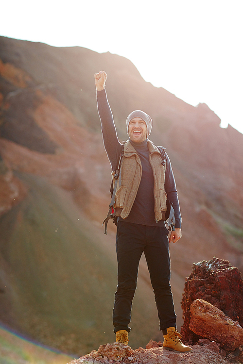 Model shot in bright sunlight of adventurous handsome traveler wearing fashionable active outwear standing on top of peak raising hand up looking at view and enjoying freedom at hike in mountains