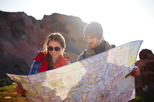 Travelling couple, young man and woman, stopping to look at map with directions during mountain hike in bright sunlight