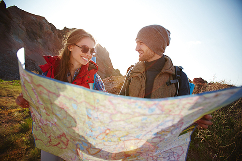 Joyful travelers, young man and woman, taking a hike through mountains in bright sunlight, stopping to look at huge map with directions
