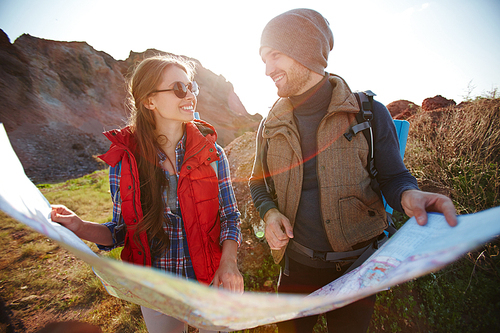 Lens flare image of smiling young couple, man and woman, looking at each other while holding big map at hike in mountains on sunny day