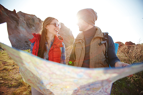 Lens flare image of joyful young tourist couple looking at each other while holding big map at hike in mountains on sunny day