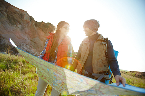 Lens flare image in bright sunlight of happy tourist couple, man and woman,  looking at each other while holding huge map on hiking path in mountains