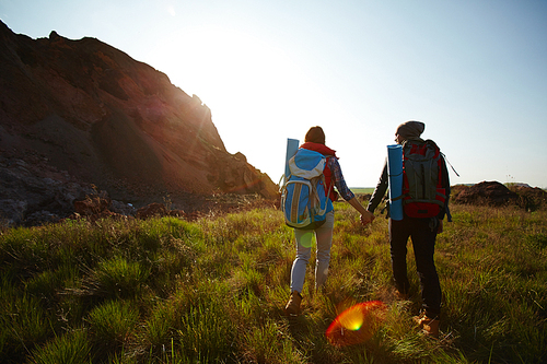 Rear view image with lens flare: young tourist couple traveling with big backpacks in nature, holding hands while walking by mountain path at sunset