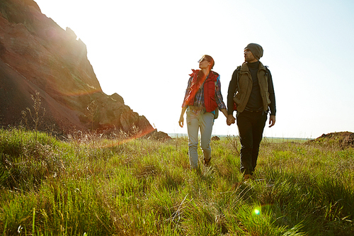 Young couple, man and woman, wearing fashionable tourist outwear, walking together holding hands during hike in mountains