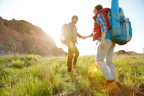 Active couple, young man and woman, traveling in mountains with big tourist backpacks, climbing uphill together on green grass, holding hands in bright sunlight