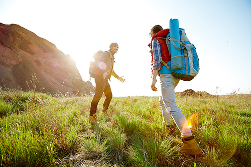 Active young couple traveling in mountains with big tourist backpacks, climbing uphill on green grass together on hiking path in bright sunlight