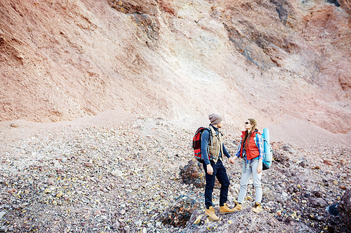 Wide shot portrait of young couple, man and woman, wearing tourist gear and big backpacks standing on hill holding hands against background of mountain on hike