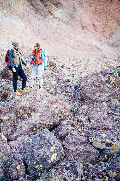 Wide shot vertical portrait of small  young couple, man and woman, wearing tourist gear and big backpacks standing on hill holding hands against background of mountain on hike