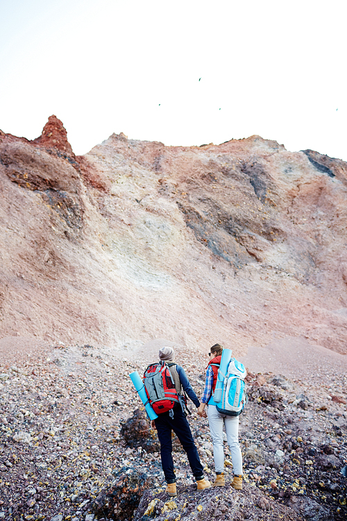 Rear view portrait of young couple, man and woman, wearing tourist gear and big backpacks standing on hill holding hands while traveling on hike in mountains