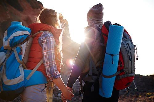 Rear view portrait of young couple, man and woman, wearing tourist gear and big backpacks standing close together holding hands against the sun, traveling in mountains on hiking path