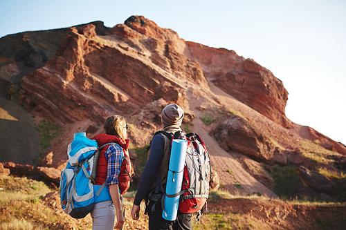 Rear view portrait of young couple, man and woman, wearing tourist gear and big backpacks standing close together looking at mountain top, traveling on hiking path in sunlight