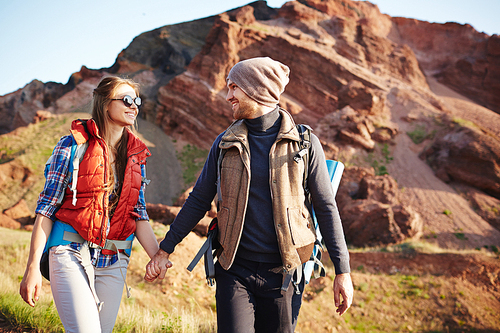 Portrait of young cheerful couple, man and woman, wearing tourist gear and big backpacks walking on hiking path through mountains holding hands, lit by bright sunlight