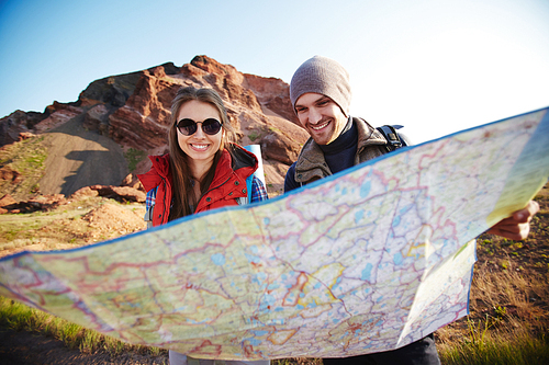 Portrait of young happy couple of adventurers holding huge map and finding their route on hiking path in mountains in bright sunlight