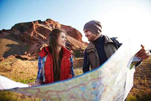 Portrait of young cheerful couple of adventurers holding huge map and and looking at each other smiling on hiking path in mountains lit by bright sunlight