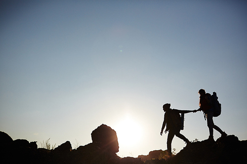 Wide shot side view image of backlit couple walking on mountain top against the sun, young woman following boyfriend holding him by hand