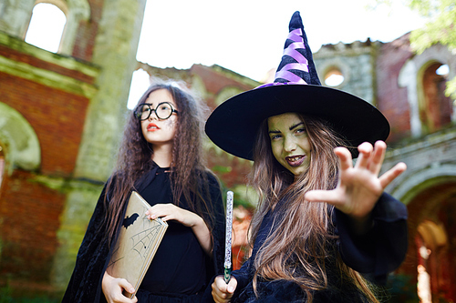 Little witch in black hat and warlock and her friend with book frightening people in abandoned place