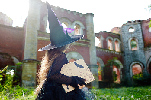 Little witch with book of spells walking towards her castle in abandoned place