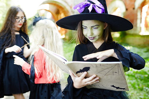 Evil girl in hat and witch attire keeping her hand over open spell-book during halloween magic