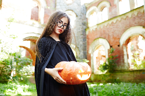 Girl in black warlock holding ripe carved pumpkin with burning candle inside