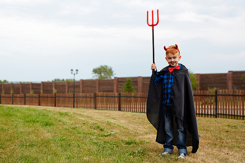 Little boy in costume of devil standing on the lawn in countryside