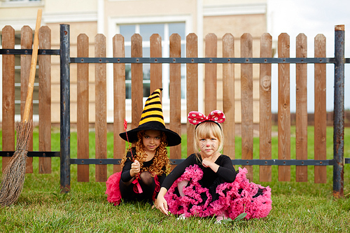 Two little girls in halloween gowns sitting on green lawn by wooden fence
