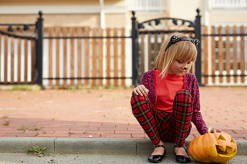 Little halloween girl with jack-o-lantern sitting on pavement not far from fence