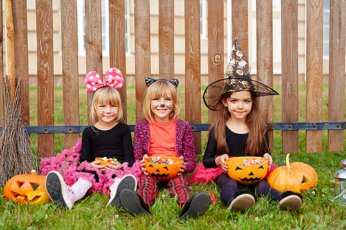 Youthful girls in halloween costumes sitting on grass by fence and eating treats