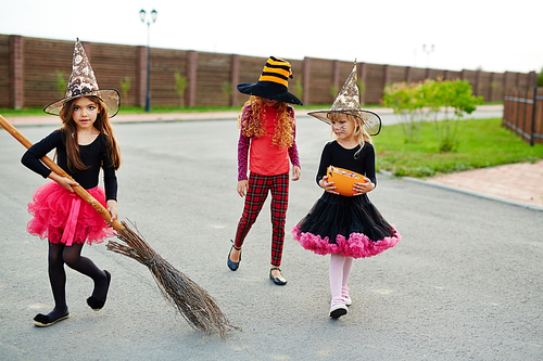 Little witch with broom and her friends walking down road on halloween evening