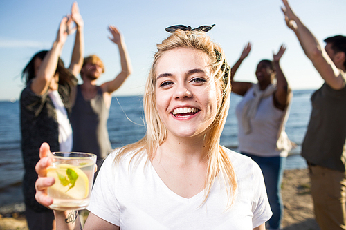 Happy girl with drink and her dancing friends on background enjoying beach party