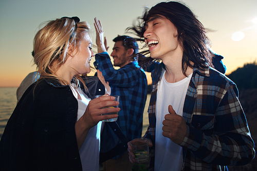 Laughing young couple with glasses of cocktails and their friends dancing at sunset on the beach