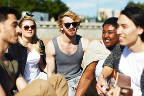 Multi-ethnic group of friends enjoying hot sunny day outdoors while handsome Asian man playing guitar, blurred background