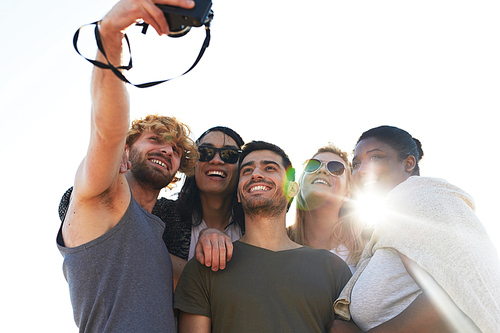 Low angle view of cheerful young friends with toothy smiles taking picture of themselves on camera while hanging out at outdoor party, lens flare