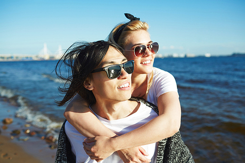 Loving young couple wearing sunglasses enjoying picturesque view while walking along seashore, blue water and cloudless sky on background