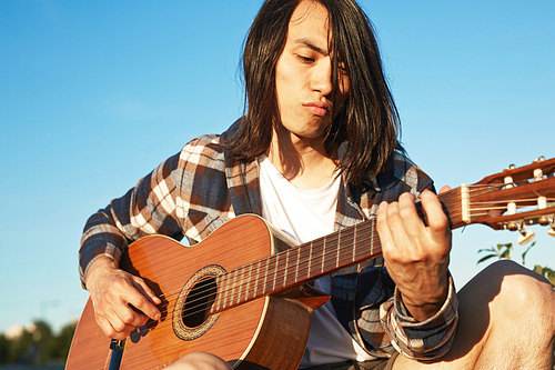 Portrait of confident Asian man wearing checked shirt playing guitar while sitting outdoors, cloudless blue sky on background