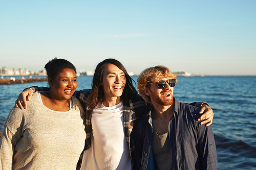 Multi-ethnic group of smiling friends enjoying picturesque view while walking along seashore, blue water surface and cloudless blue sky on background