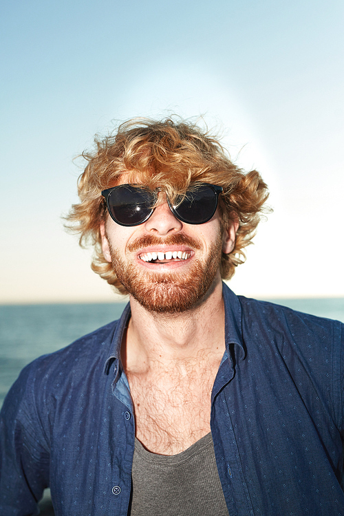 Head and shoulders portrait of handsome bearded man wearing sunglasses  with toothy smile while enjoying sunny day on seashore