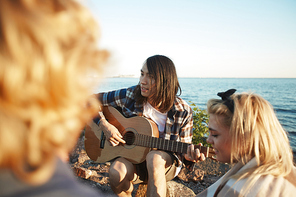 Group of young friends relaxing around campfire and listening to music, handsome Asian man playing guitar, picturesque seascape on background